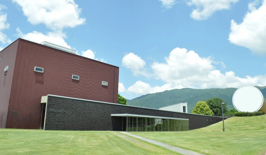 芝生の広場に建つ美術館の外観。赤茶色の壁と黒い低層棟、奥に山並み